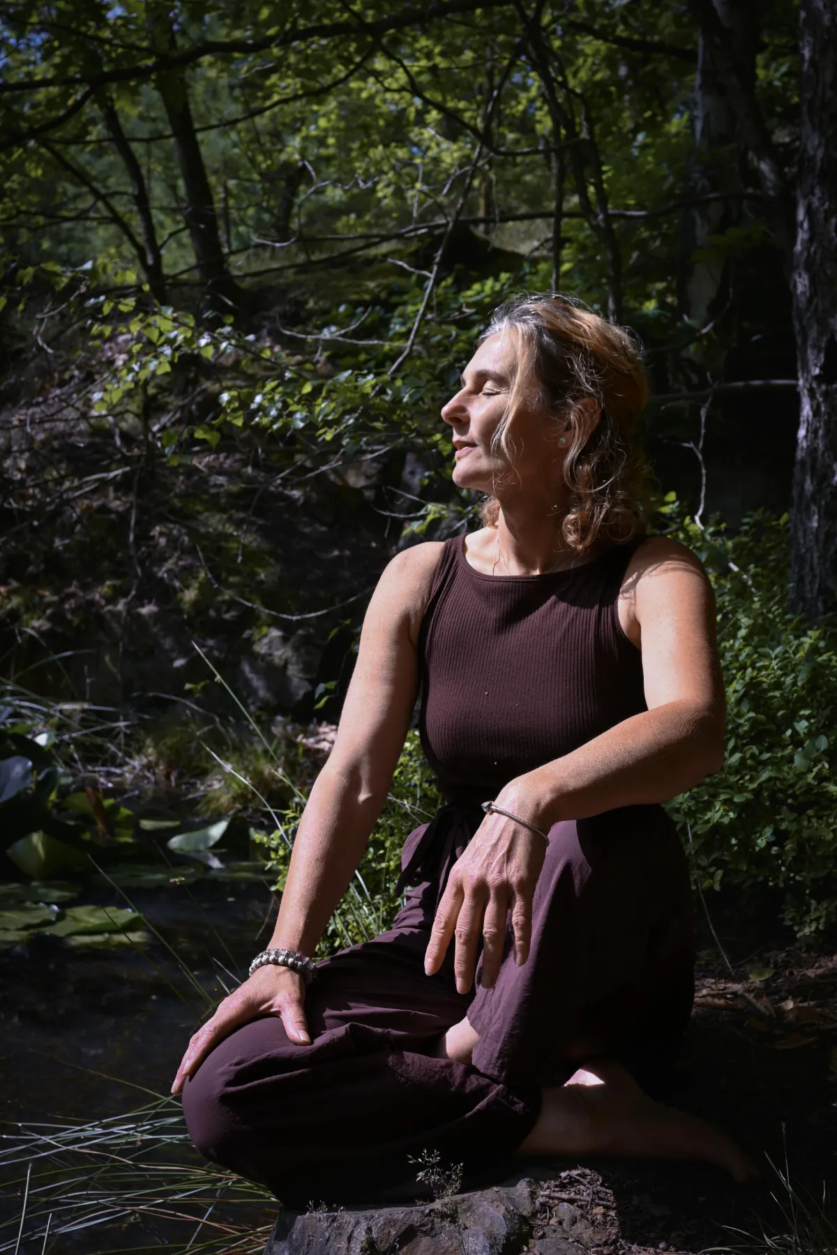 Porträt Martina Schneider-Just. Sie sitzt im Wald auf dem Boden, das Gesicht der Sonne zugewandt, die Augen geschlossen.