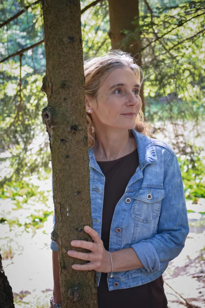 Porträt Martina Schneider-Just in Dreiviertel-Darstellung. Sie trägt eine Jeansjacke und wird teilweise von einem Baum verdeckt.