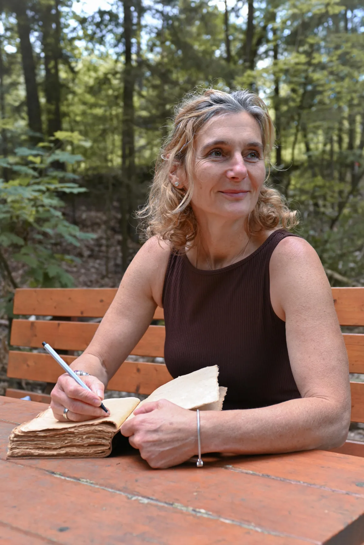 Porträt Martina Schneider-Just. Sie sitzt auf einer Bank im Wald. Auf dem Tisch vor ihr liegt ein Buch, in dem sie sich Notizen macht.