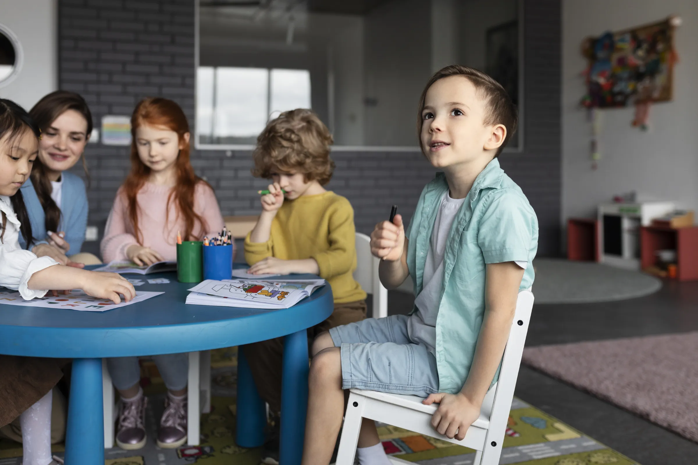 Eine Kindergartengruppe sitzt mit Erzieherin an einem Tisch. Im Vordergrund ist ein lächelnder Junge im Fokus