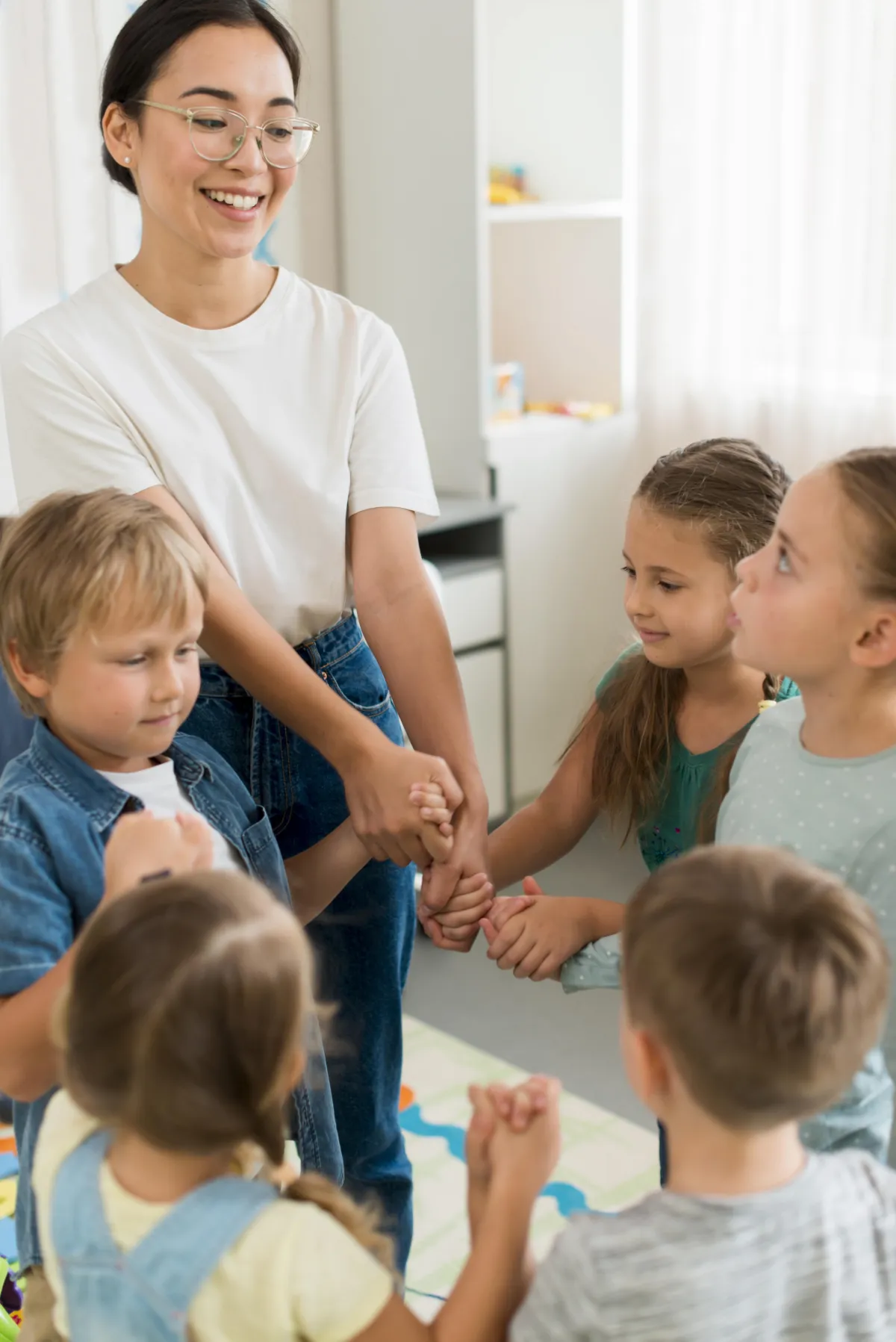 Eine Grundschullehrerin spielt mit ihren Schülern und Schülerinnen. Alle halten sich an den Händen und haben einen Kreis gebildet.
