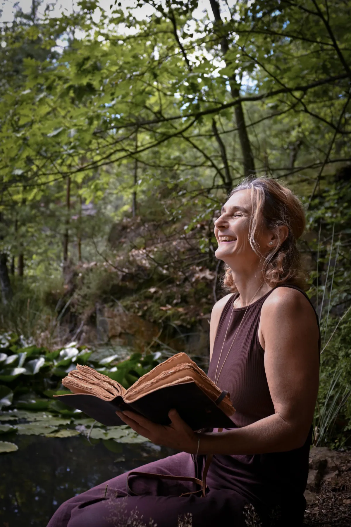 Porträt Martina Schneider-Just. Sie sitzt an einem Gewässer im Wald. Sie hält ein aufgeschlagenes Buch und lacht.