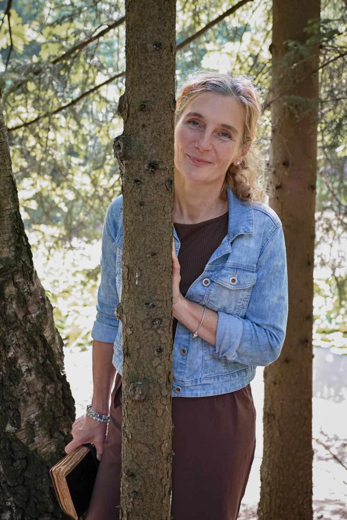 Dreiviertelporträt Martina Schneider-Just. Sie steht hinter einem dünnen Baum und schaut direkt in die Kamera. In der rechten Hand hält sie ein Buch.